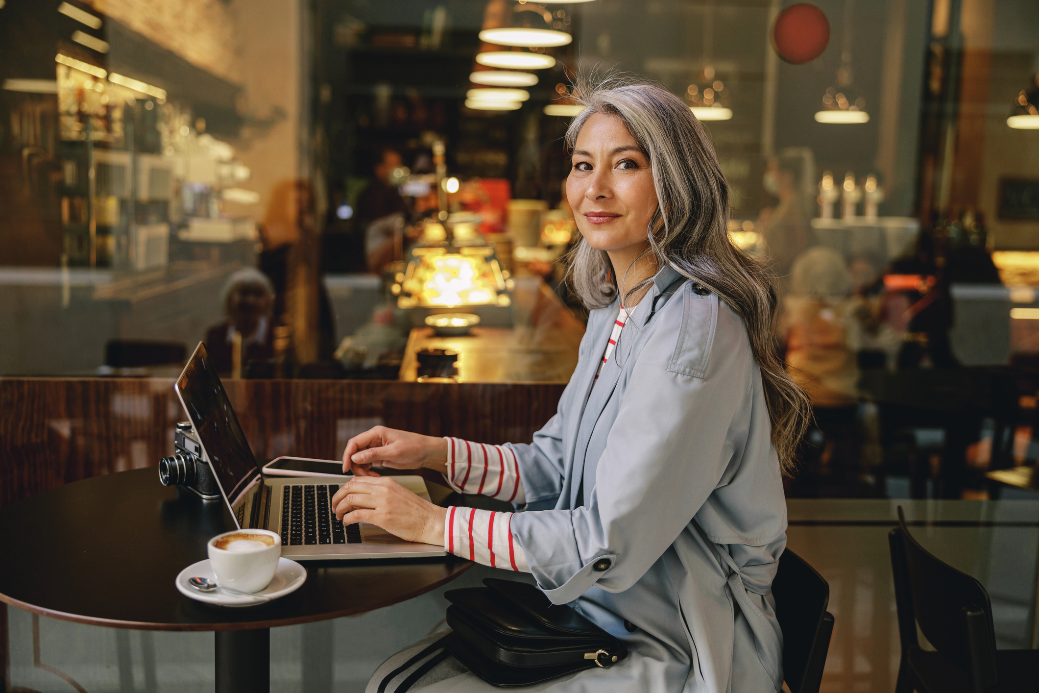 Attractive mature woman in trench coat working on laptop with coffee cup on table in modern cafe